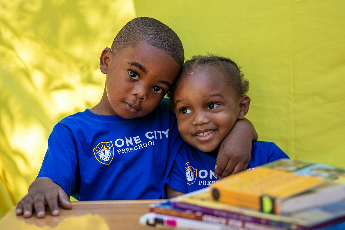 Two children wearing blue "One City Preschool" shirts sit closely, with one child hugging the other next to a stack of books, against a yellow background
